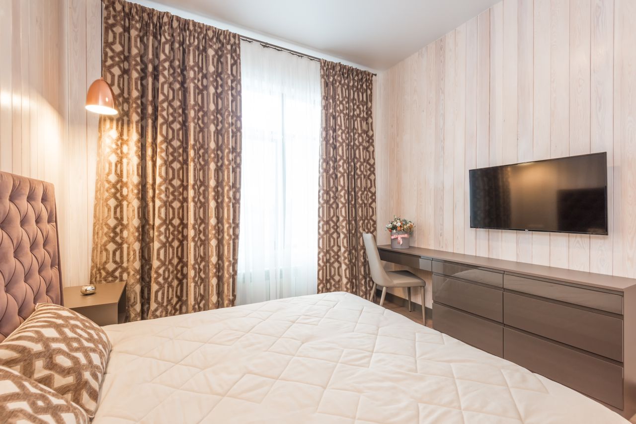 Comfortable bed with soft cushions and headboard in classic styled bedroom with modern TV set and chair